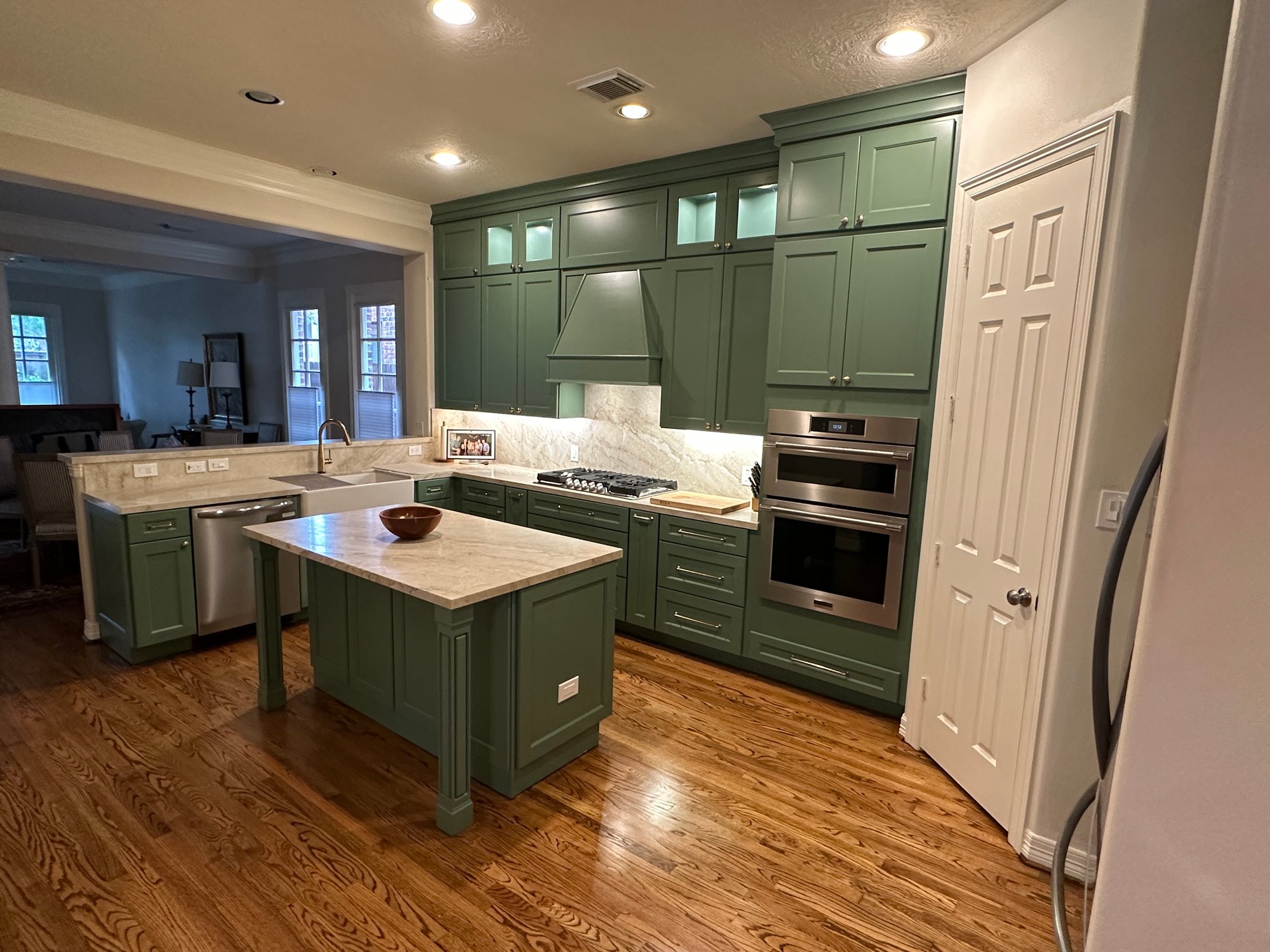 Renovated kitchen in a home in Houston, Texas, with sage green shaker cabinets, white marble countertops, a central island, farmhouse sink, and stainless steel double wall oven and gas cooktop.
