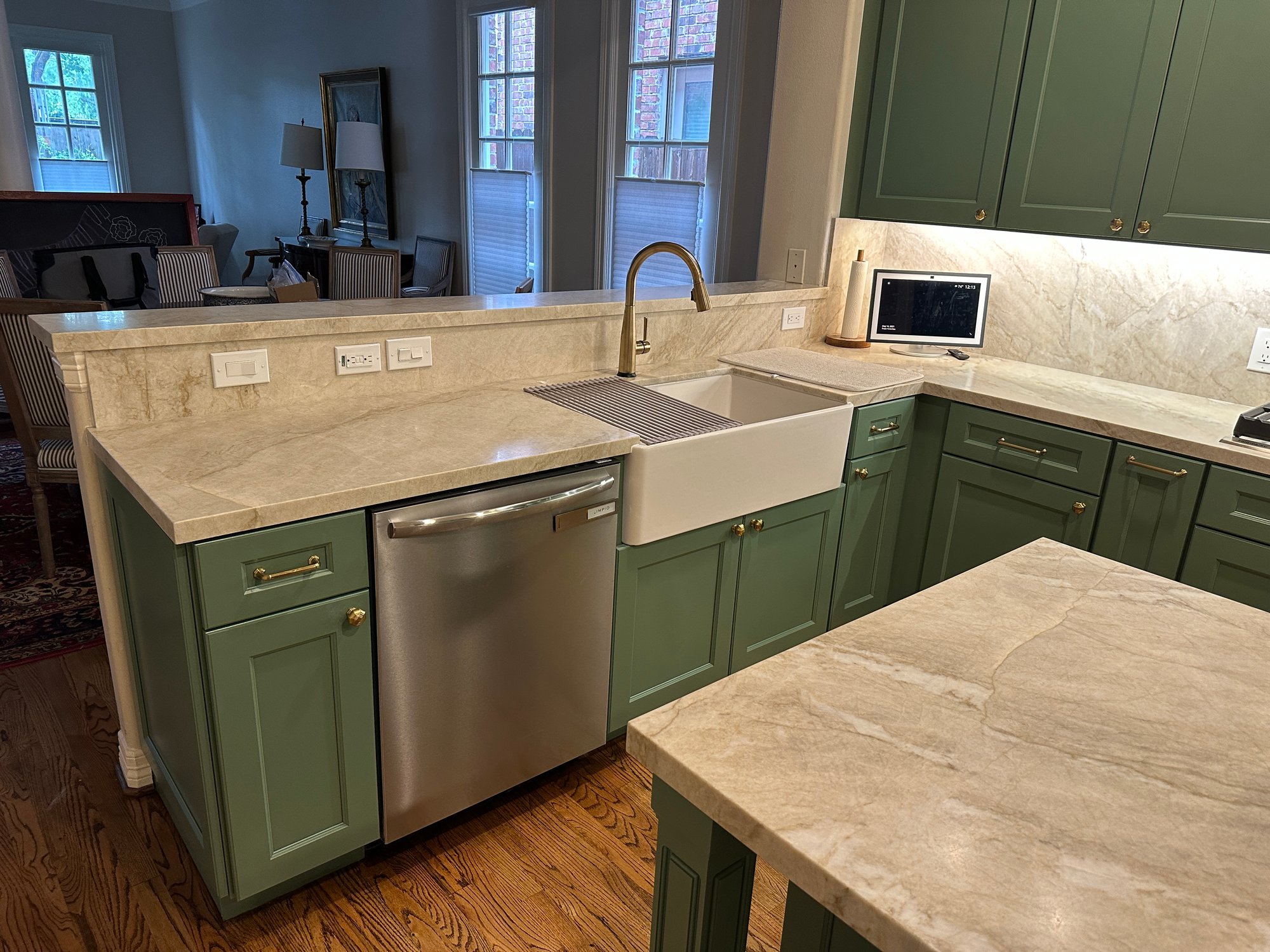 Kitchen renovation in a Houston, Texas, home with farmhouse sink, gold faucet, stainless steel dishwasher, sage green cabinets, and marble countertops, with an adjoining sitting room in view.