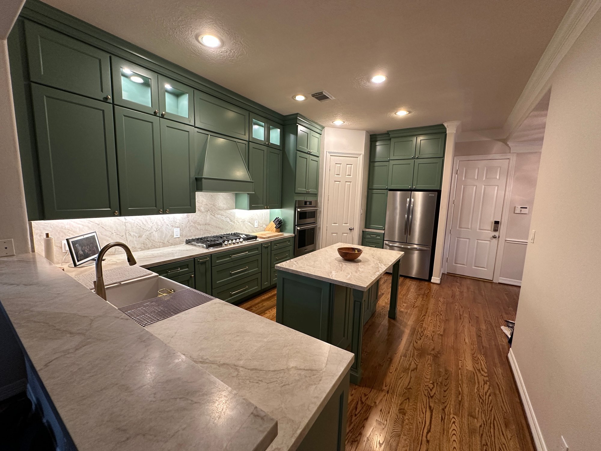 Renovated kitchen in a Houston, Texas, home with sage green cabinets, marble countertops, farmhouse sink, gas cooktop, built-in refrigerator, and center island, viewed from the peninsula.