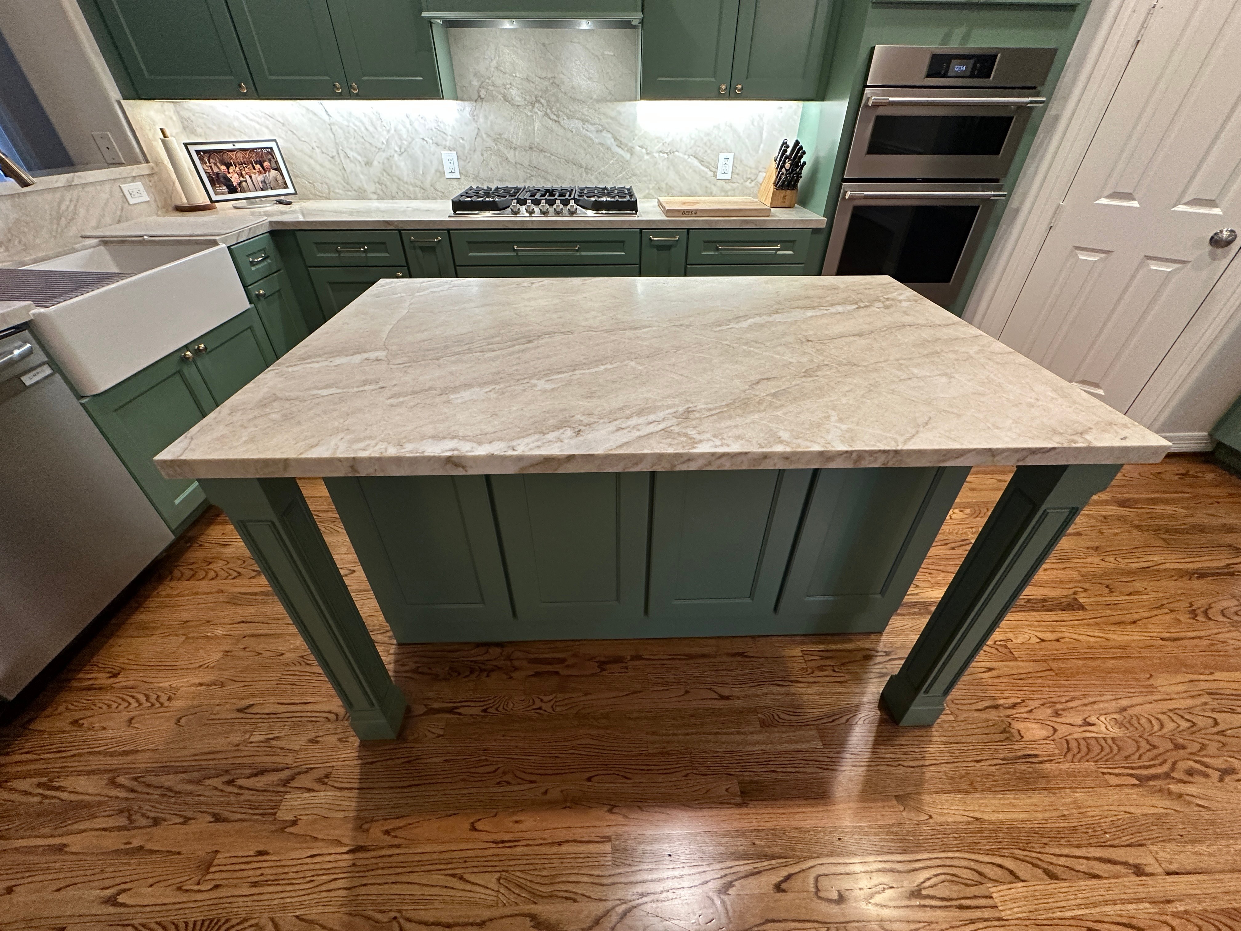 Kitchen island inside a Houston, Texas, home with marble waterfall-style countertop and turned wood legs, sage green cabinets, gas cooktop, and double wall oven in the background.