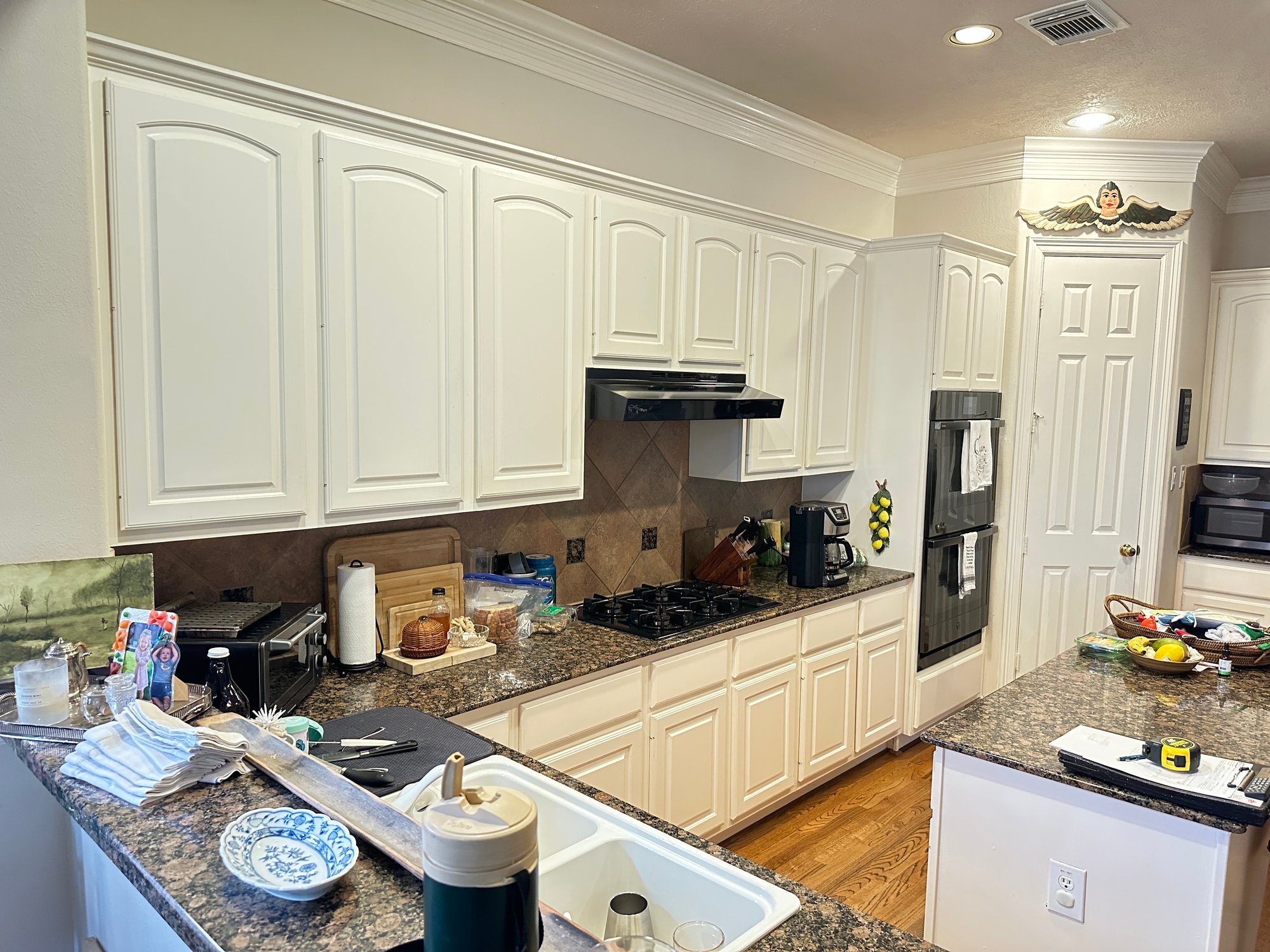 Original kitchen in a home in Houston, Texas, before renovation with white raised-panel cabinets, dark speckled granite countertops, tile backsplash, and black double wall oven.