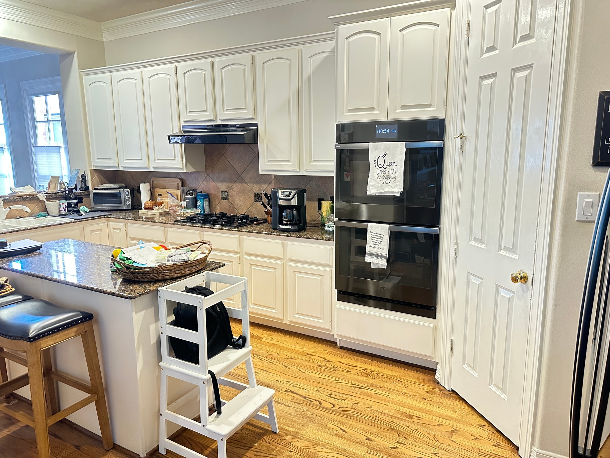 Original kitchen in a Houston, Texas, home before renovation with white raised-panel cabinets, dark granite countertops, tile backsplash, and black double wall oven with an angled peninsula island.