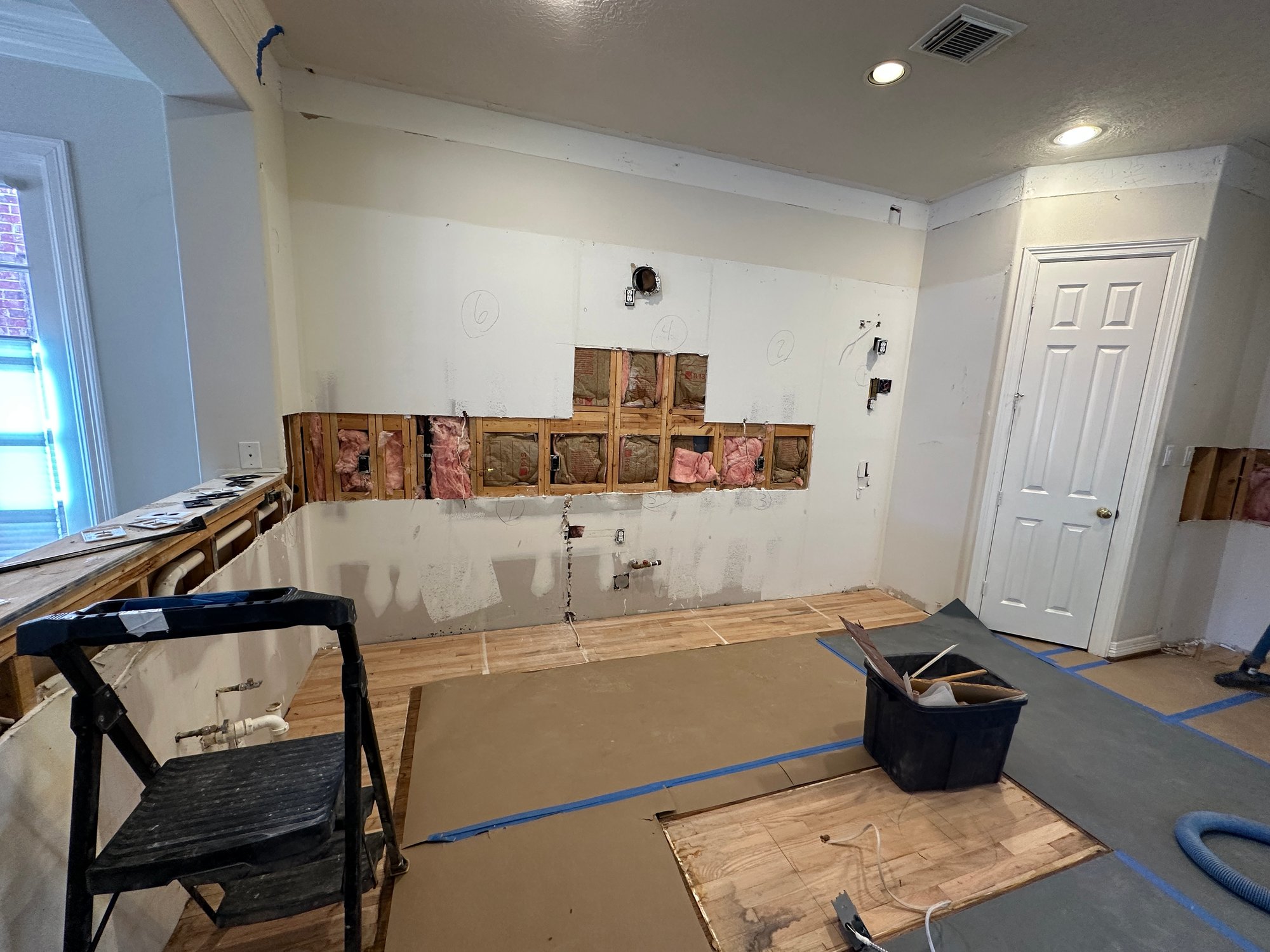 Kitchen of a Houston, Texas, home in mid-renovation with cabinets and countertops removed, exposed wall studs and insulation, and plumbing rough-in visible.